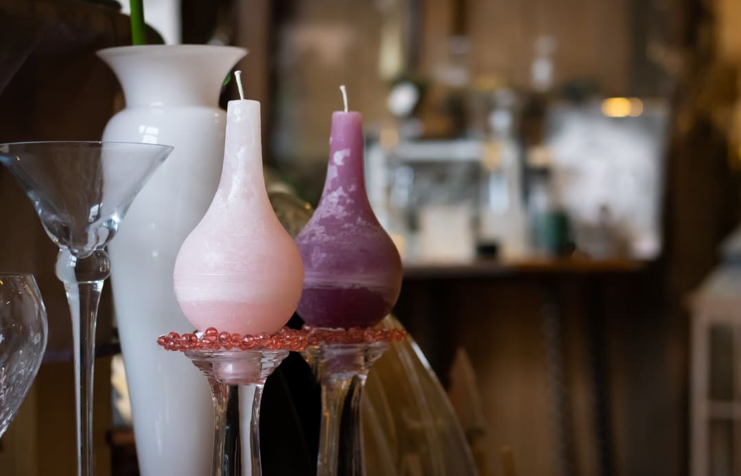 Zwei bauchige Kerzen in Rosa und Pflaume auf Glasständern vor einer weißen Vase