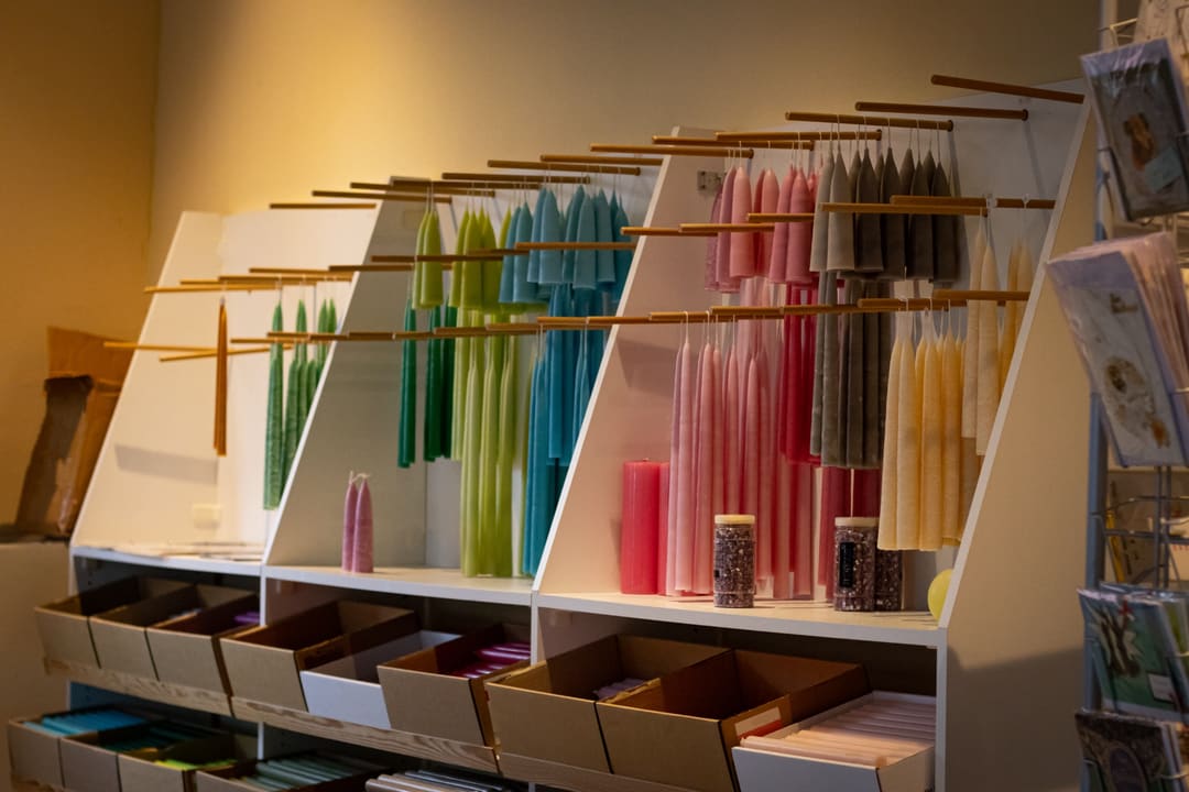 Gezogene Stabkerzen hängen paarweise an Holzstäben, sortiert nach Farben von Grün über Blau bis Rosa